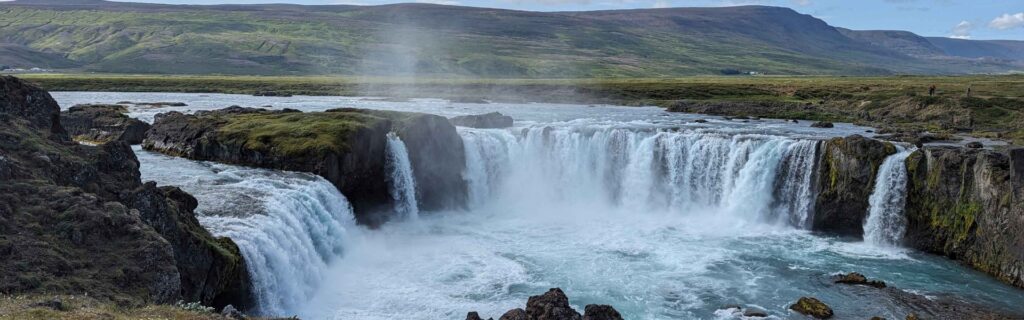 Panoramic view of Goðafoss waterfall with green hills and blue sky