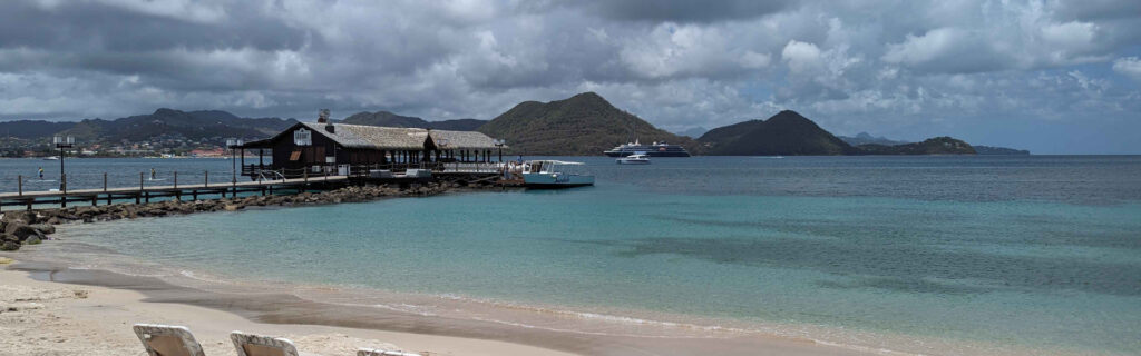 A panoramic beach view with turquoise water a rustic pier and cruise ships in the distance