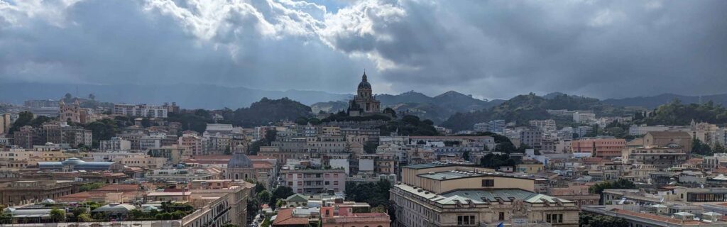 A panoramic view of Messina shows city buildings and a domed church under dramatic clouds