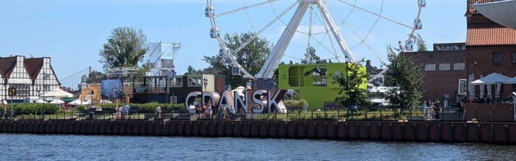 Gdańsk sign and Ferris wheel along the Motława River during the St. Dominic's Fair under the midday summer sun