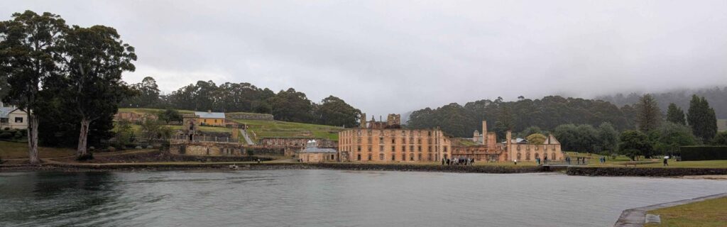 The Penitentiary ruins at Port Arthur Historic Site stretch across the waterfront beneath misty wooded hills.